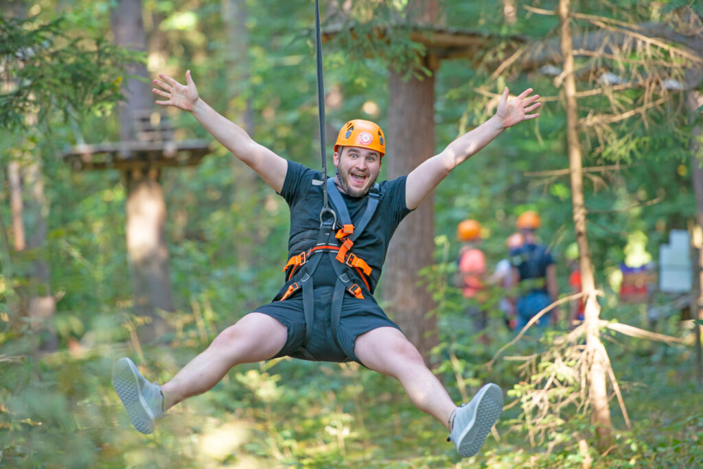 Ein Mann gleitet einen Flying Fox im Kletterwald Vaterstetten hinunter, streckt dabei die Arme und Beine vom Körper und Lacht direkt in die Kamera. Im Hintergrund sind weitere besucher, diese befinden sich am Bden.