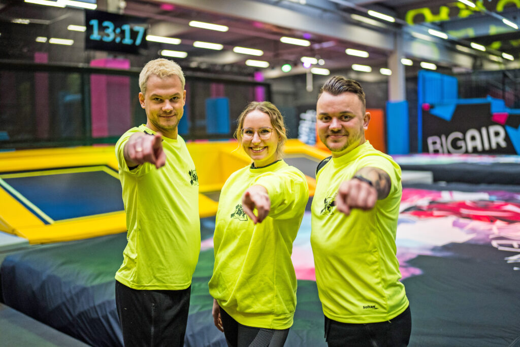 Drei Personen, welche gelbe Mitarbeiterkleidung der MAXX Arena tragen. Im Hintergrund ist der Indoor Freizeitpark MAXX Arena zu sehen. Die Personen zeigen mit den Fingern in die Kamera.