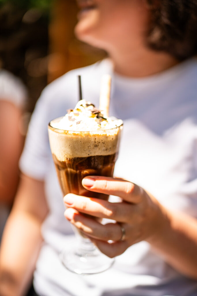 Im Fokus ist ein kühler Eiskaffee, welcher mit Waffel und Strohhalm serviert wird. Im Hintergrund ist eine Frau, welche das Glas in der Hand hält. Sie sitzt bei der Fater Alm im Münchner Wald.