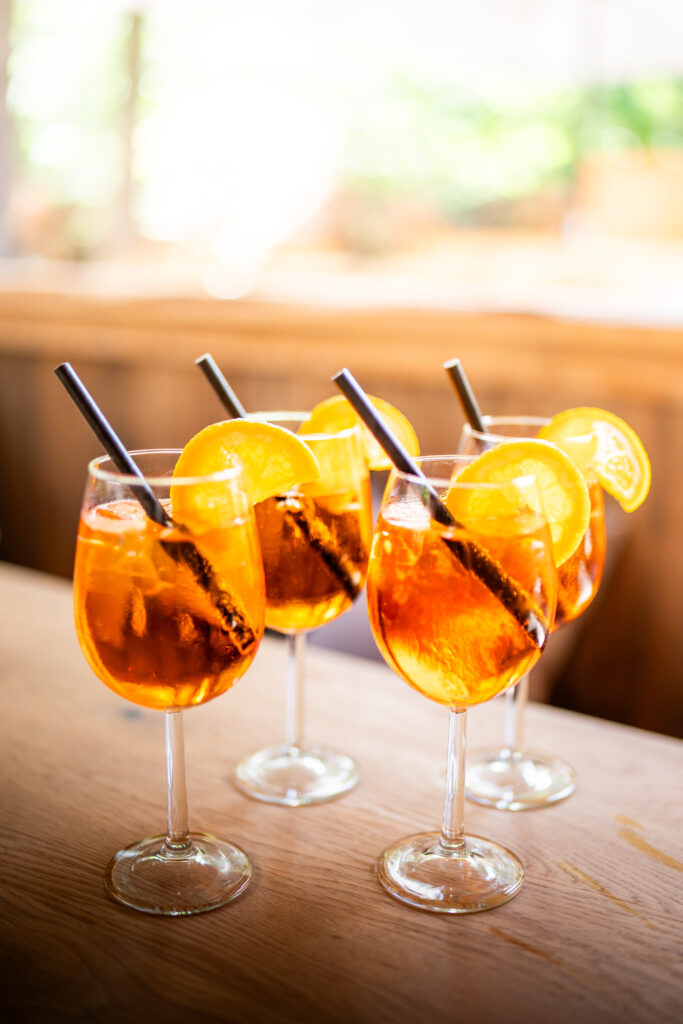 Fünft Gläser Aperol Spritz, mit schwarzem Strohhalm und Orange im Glas. Sie stehen auf einem braunen Holztisch.