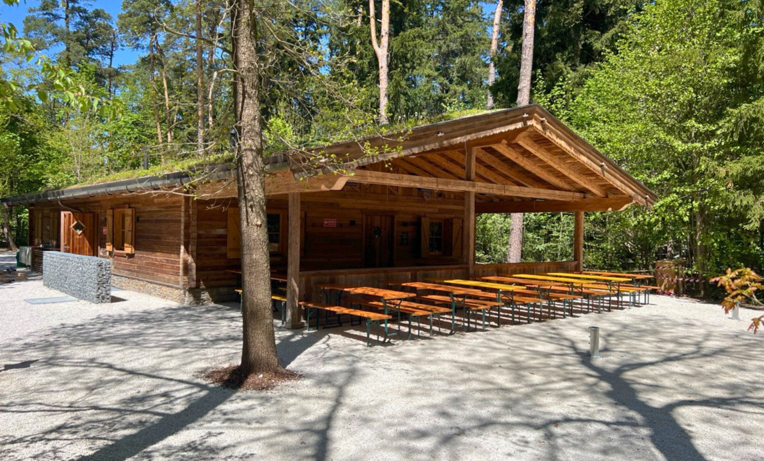 Bayerische Berghütte zwischen den Bäumen im Wald, mit Biertischen und viel Platz davor.