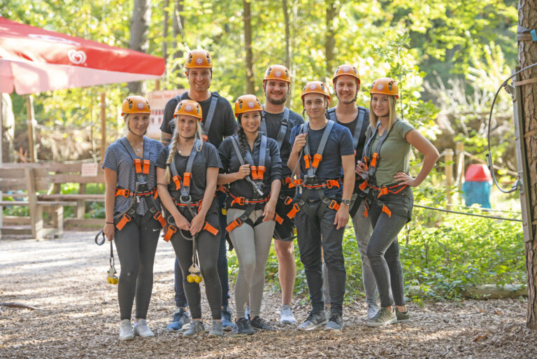 Ein Gruppenfoto von vier Männern und vier Frauen, welche im Kletterwald Vaterstetten stehen und in die Kamera lächeln. Sie tragen alle einen orangenen Helm und einen Kelttergurt.