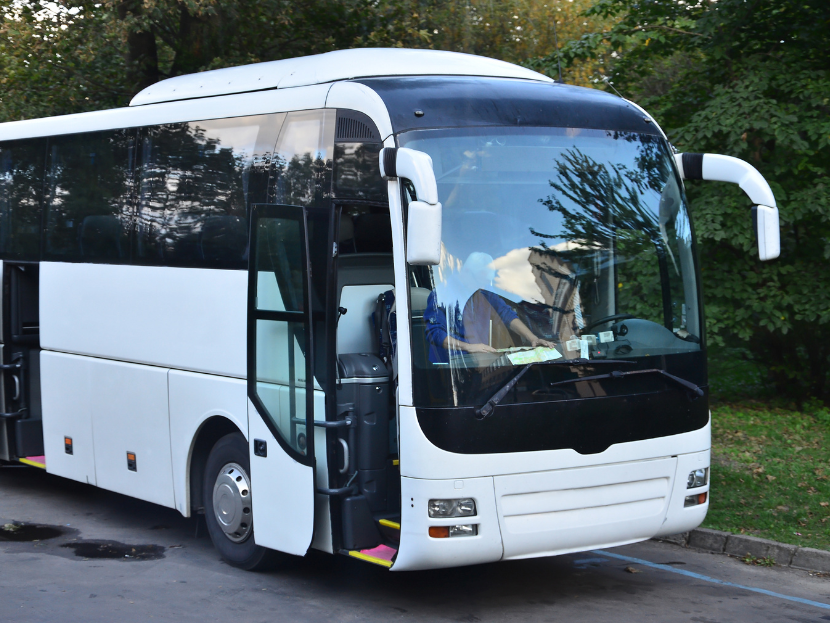 Ein weißer Bus, bei welchem die Türen offen sind da dieser auf einer Straße steht. im Hintergrund sind grüne Bäume.