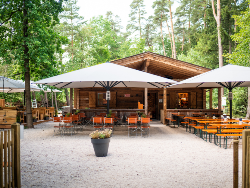 Die Fater Alm im Müncher Wald in der vollen Perspektive von vorne. Mit Waldgarten, Sonnenschirmen, links der VIP Bereich und rechts Bierbänke. Dahinter ist der Wald zu sehen.