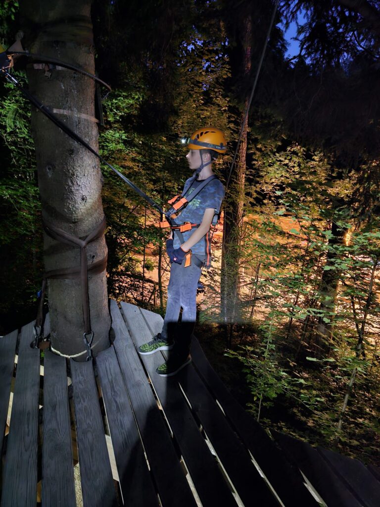 Ein Junge mit einer Stirnlampe auf dem Helm steht auf einer Plattform im Kletterwald Vaterstetten und lehnt sich nach hinten in seinen Klettergurt, sodass das sicherungsseil straff gespannt ist.