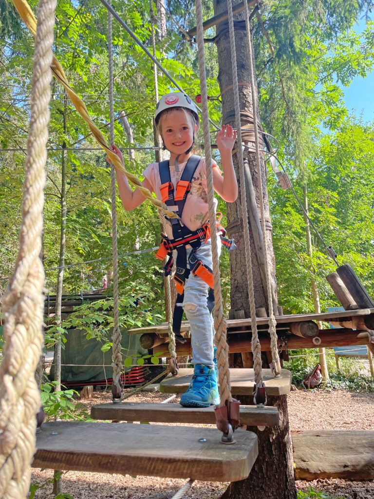 Ein Junges Mädchen klettert einen Kinderparcours im Kletterwald Vaterstetten und lacht diekt in die Kamera.