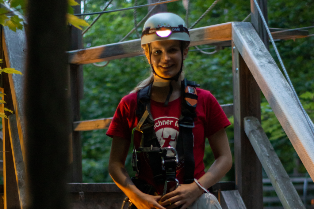 Ein junges Mächen lächelt in die Kamera, sie trägt Klettergurt und Helm, da sie im Kletterwald Vaterstetten ist. Die Atmosphäre ist dunkel, da sie beim Nachtklettern ist.