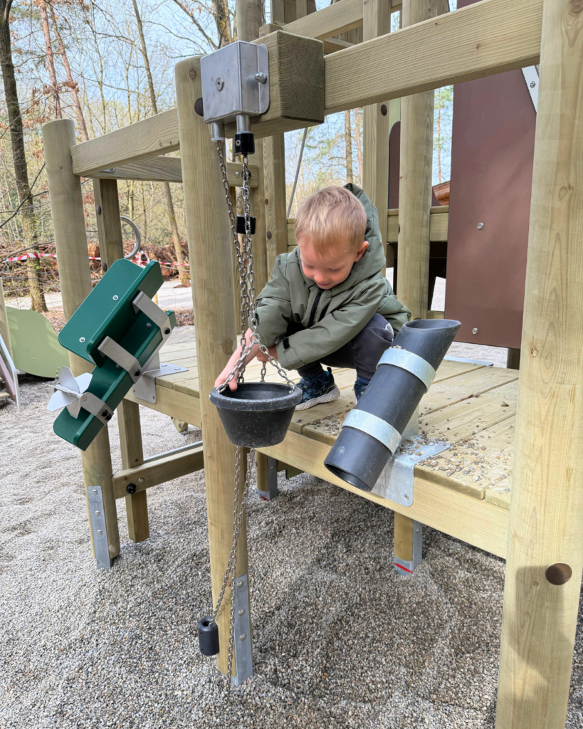 Ein Kleinkind welches auf einem Pielplatz Kieselsteine in einen Eimer füllt, welcher an einer Kette hängt. Das Kind befindet sich auf einer Plattform des Spielplatzes im Münchner Wald.